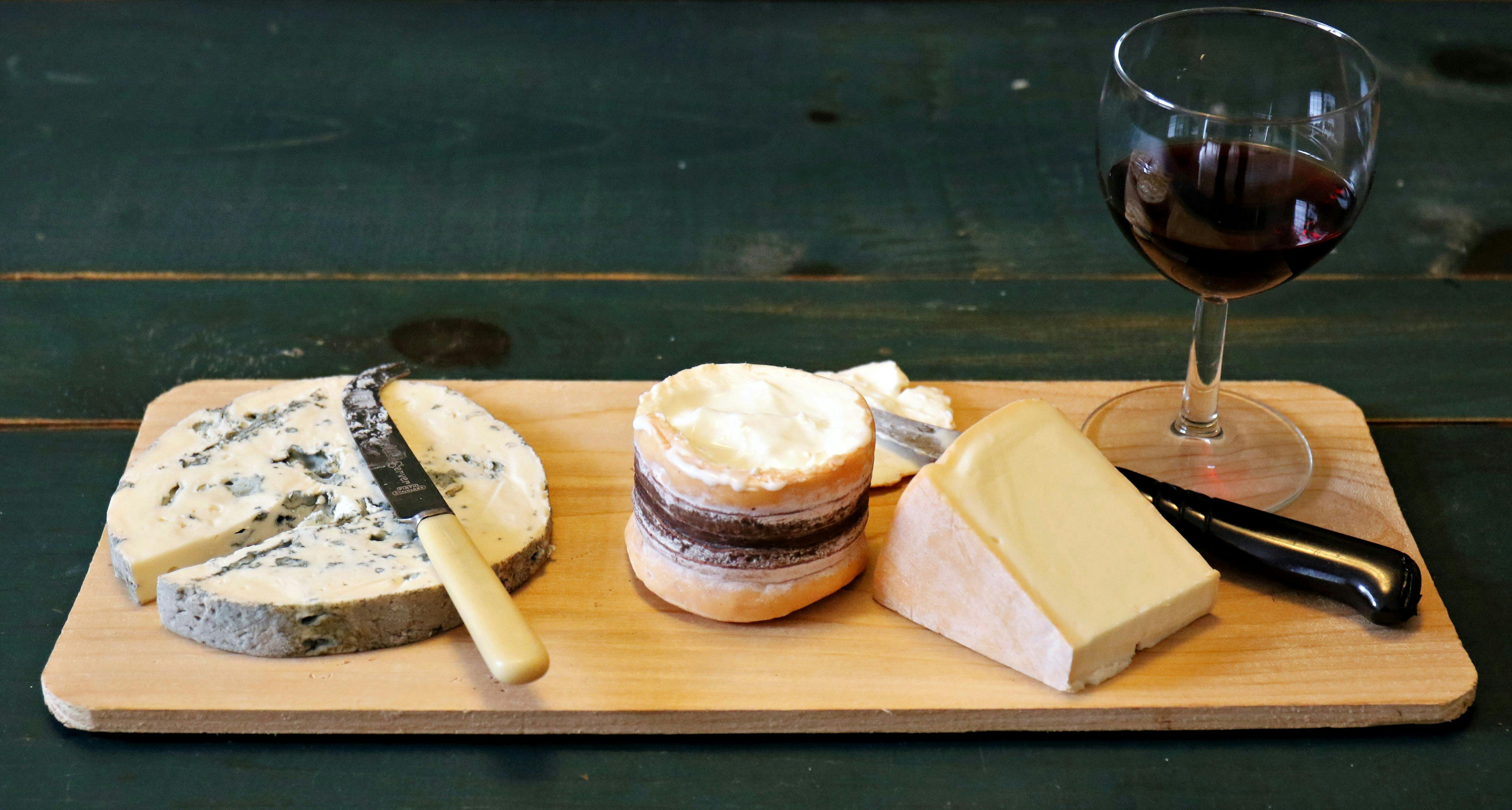 A cheeseboard with three types of cheese, knives, and a glass of red wine on a wooden surface.