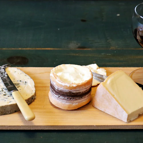 En ostbräda med tre typer av ost, knivar och ett glas rött vin på en träyta.