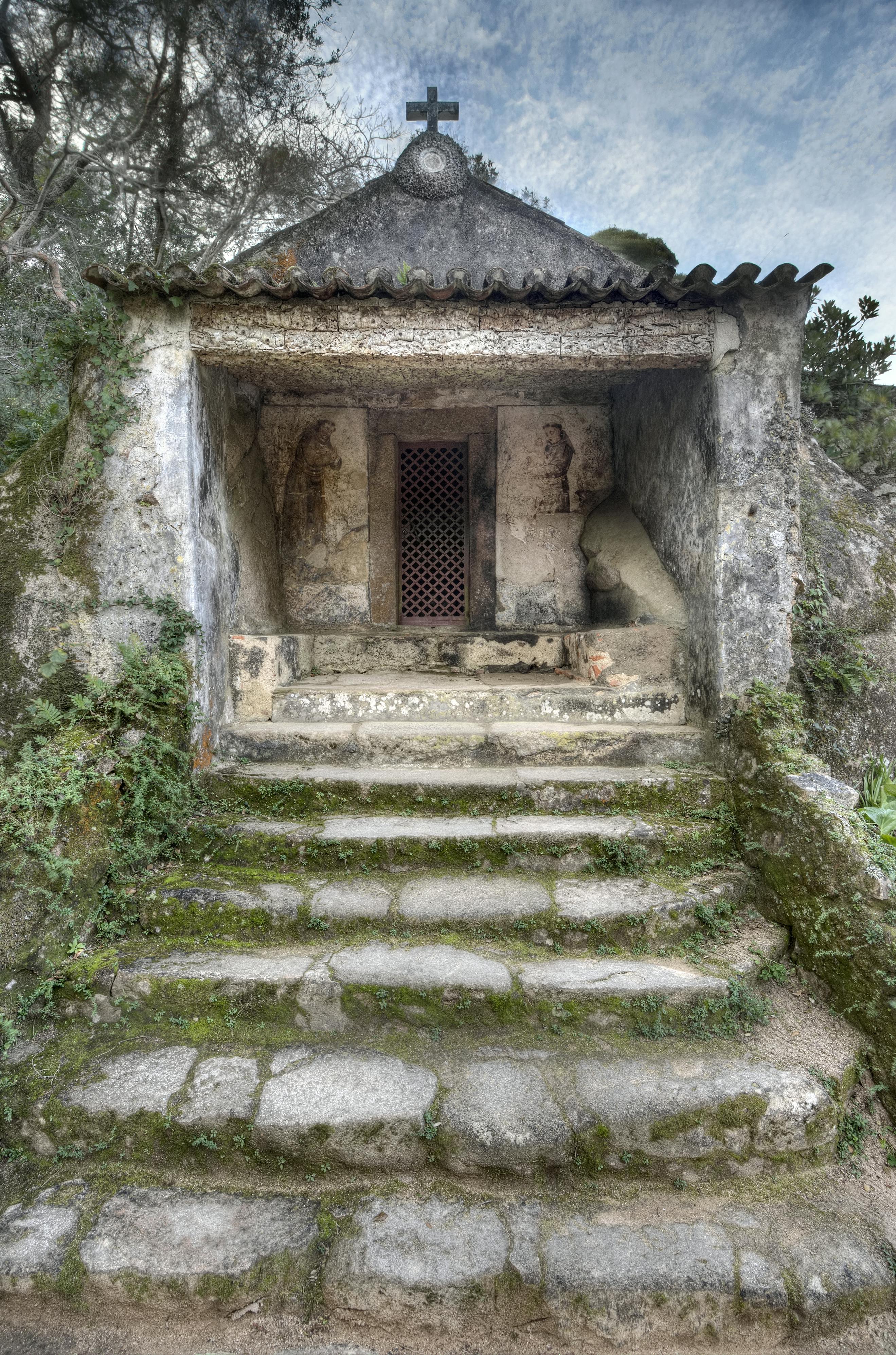 Scalini di pietra che conducono a una piccola struttura in pietra, danneggiata dalle intemperie, con antiche incisioni sul muro e una porta a grata. Intorno cresce della vegetazione.