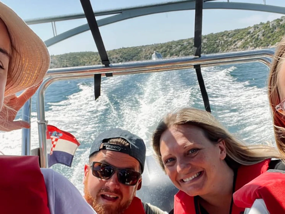 Four people smiling on a boat wearing red life vests, with the boat speeding on water under a sunny sky.