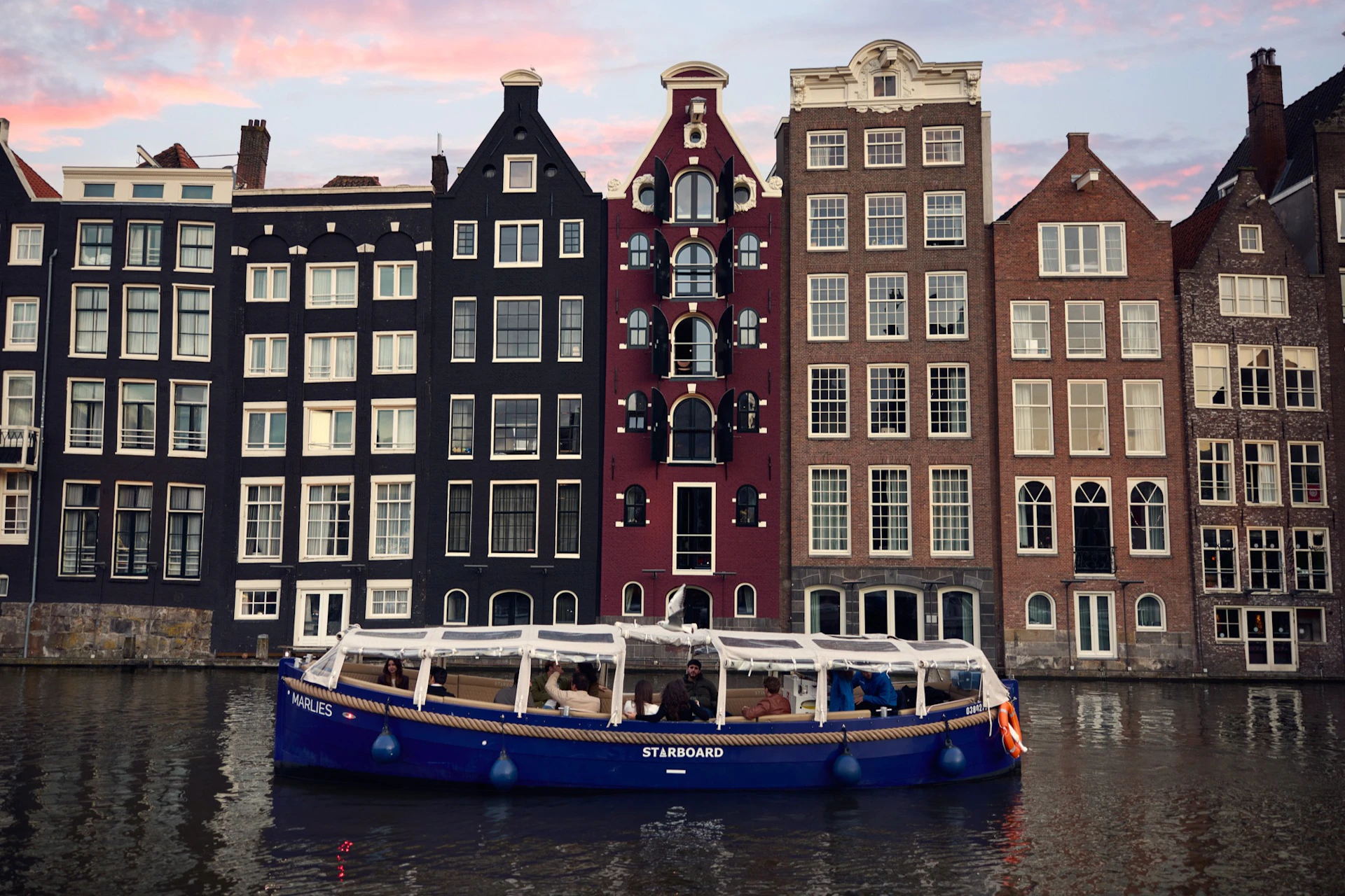A blue tour boat on a canal with historic, narrow buildings in various colors and styles in the background.