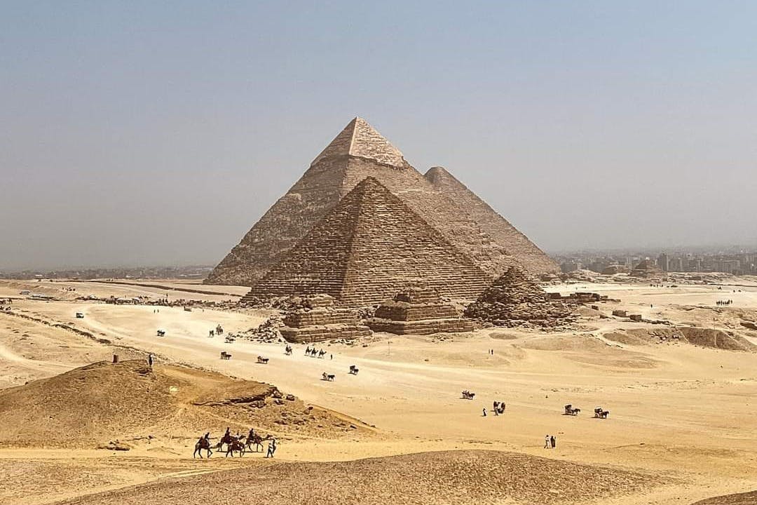 Tre pyramider i ett ökenlandskap med spridda turister, kameler och hästdragna vagnar i förgrunden. Stadssilhuetten i fjärran.