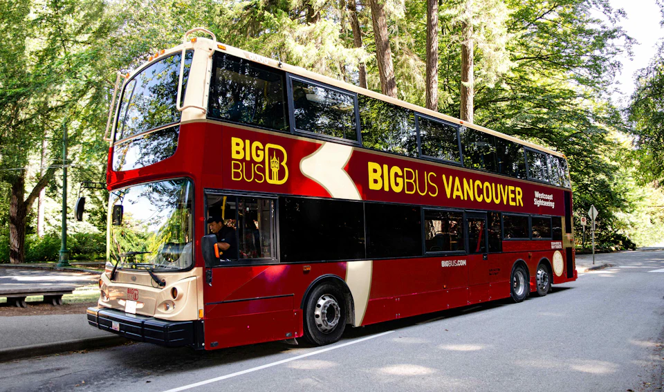 A red double-decker bus labeled "Big Bus Vancouver" is parked on a road surrounded by tall trees.