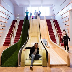 Människor som glider ner för intilliggande rutschkanor bredvid trappor i ett ljust rum med hyllor som visar cirkulära föremål på väggarna.