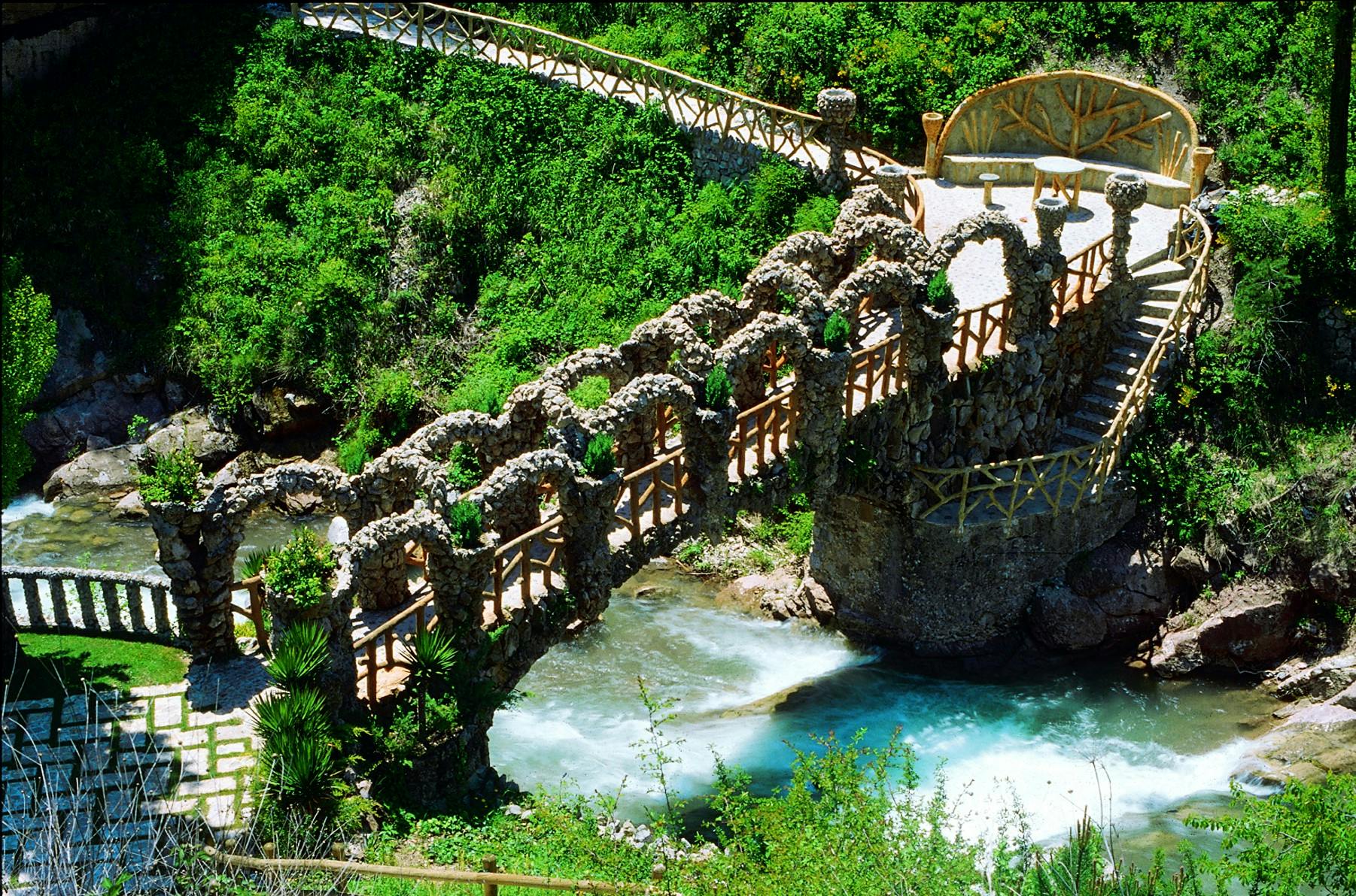 緑豊かな葉と岩場に囲まれた、アーチ型の素朴な石造りの橋が川を渡っています。