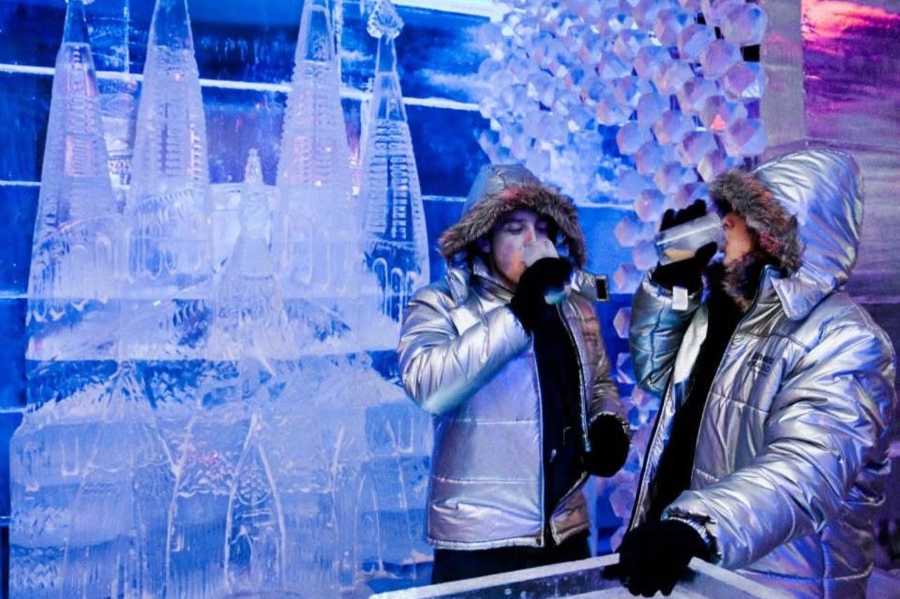 Due individui che indossano giacche invernali riflettenti e cappucci foderati di pelliccia si trovano in un bar di ghiaccio, circondati da sculture e decorazioni di ghiaccio.