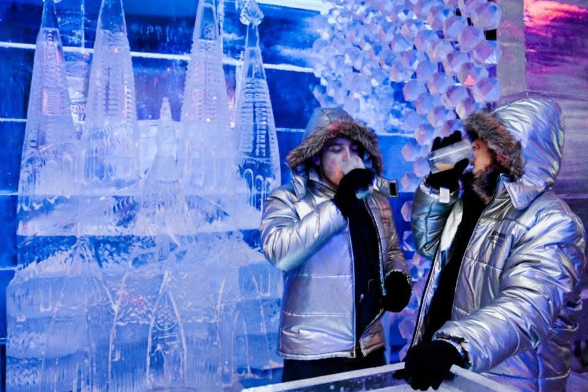 Two individuals wearing reflective winter jackets and fur-lined hoods stand in an ice bar, surrounded by ice sculptures and decor.