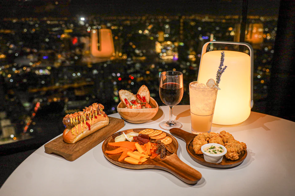 A table with various dishes including a hot dog, cheese platter, fried chicken, and drinks, set against a city nightscape.