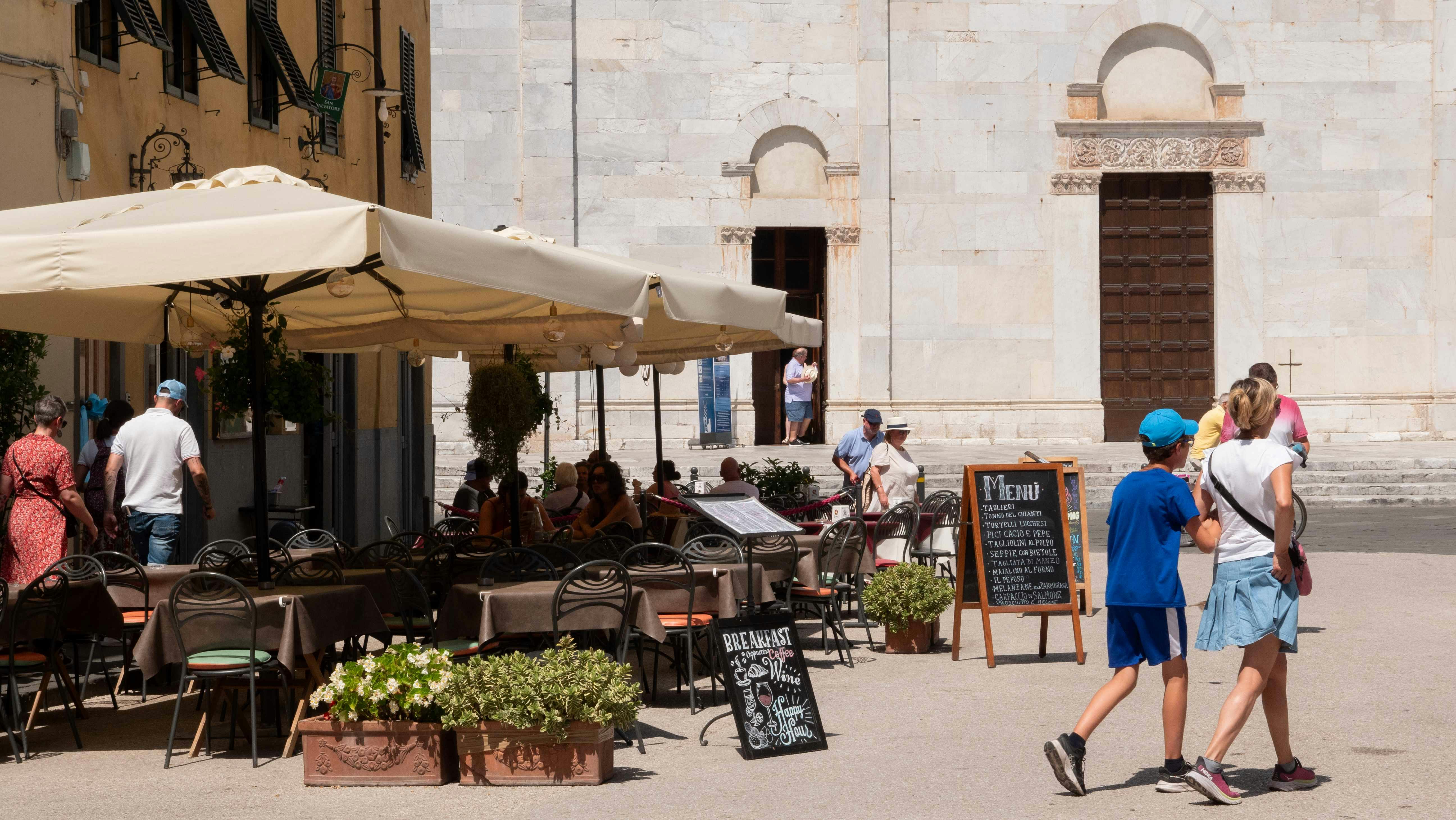 People walking in front of an outdoor café with an ornate menu board and a large building with arches and a wooden door in the background.