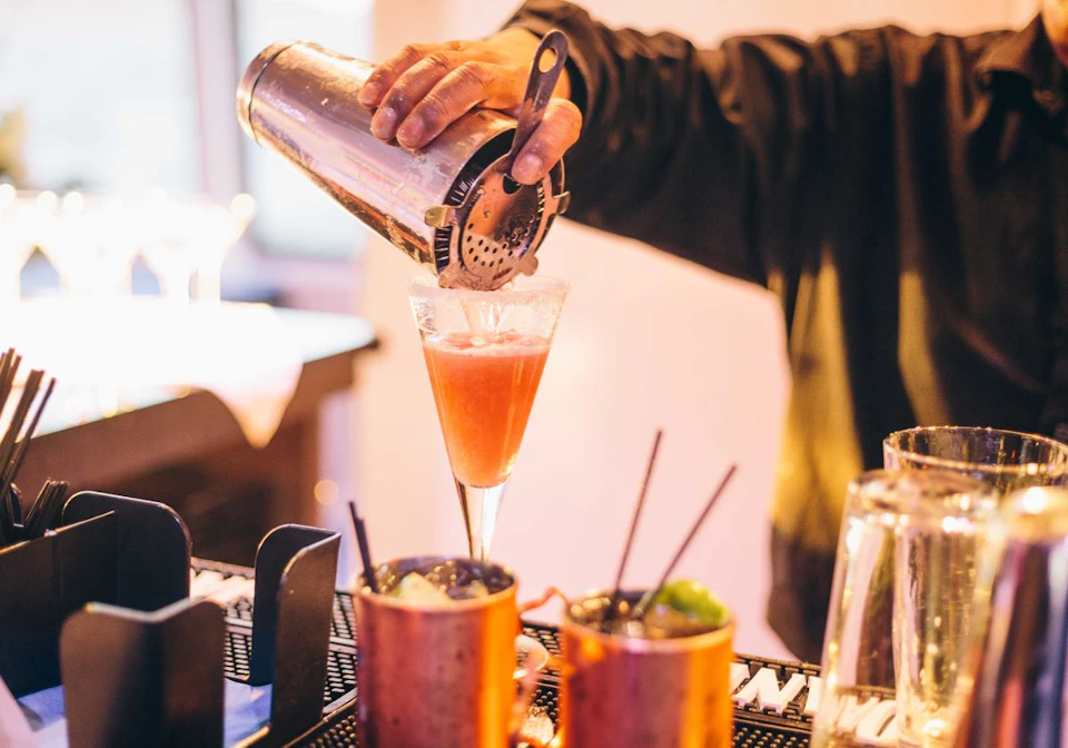 Person pouring a pink cocktail from a shaker into a glass, with two copper mugs and a bar in the background.