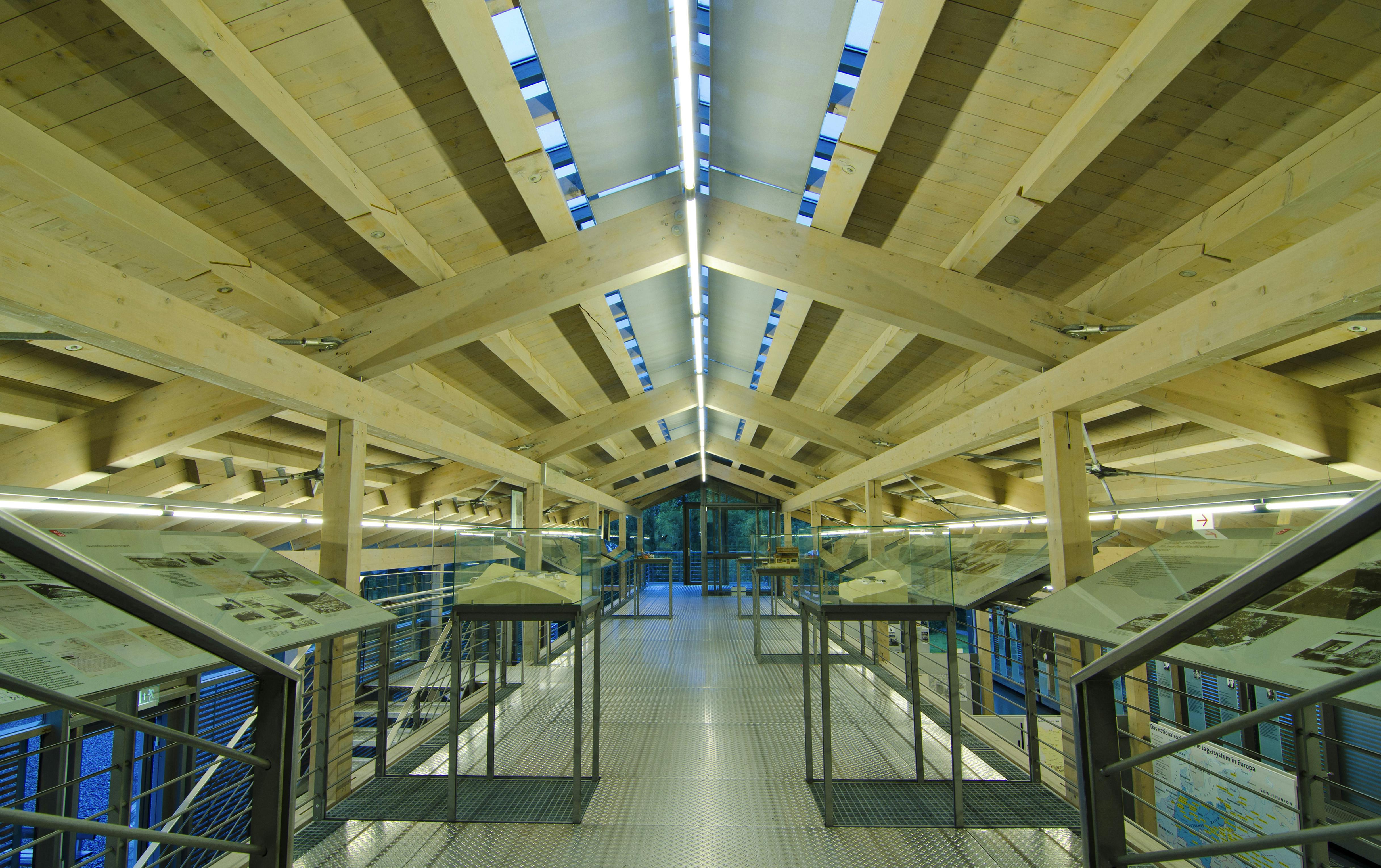 A modern exhibition hall with wooden beams and a metal walkway, displaying various historical artifacts on raised platforms.
