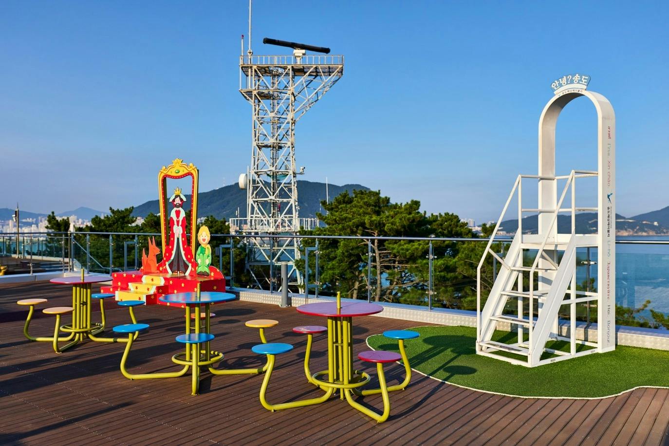 A colorful rooftop with children's play equipment, tables, a radar tower, and a scenic view with trees and a blue sky.