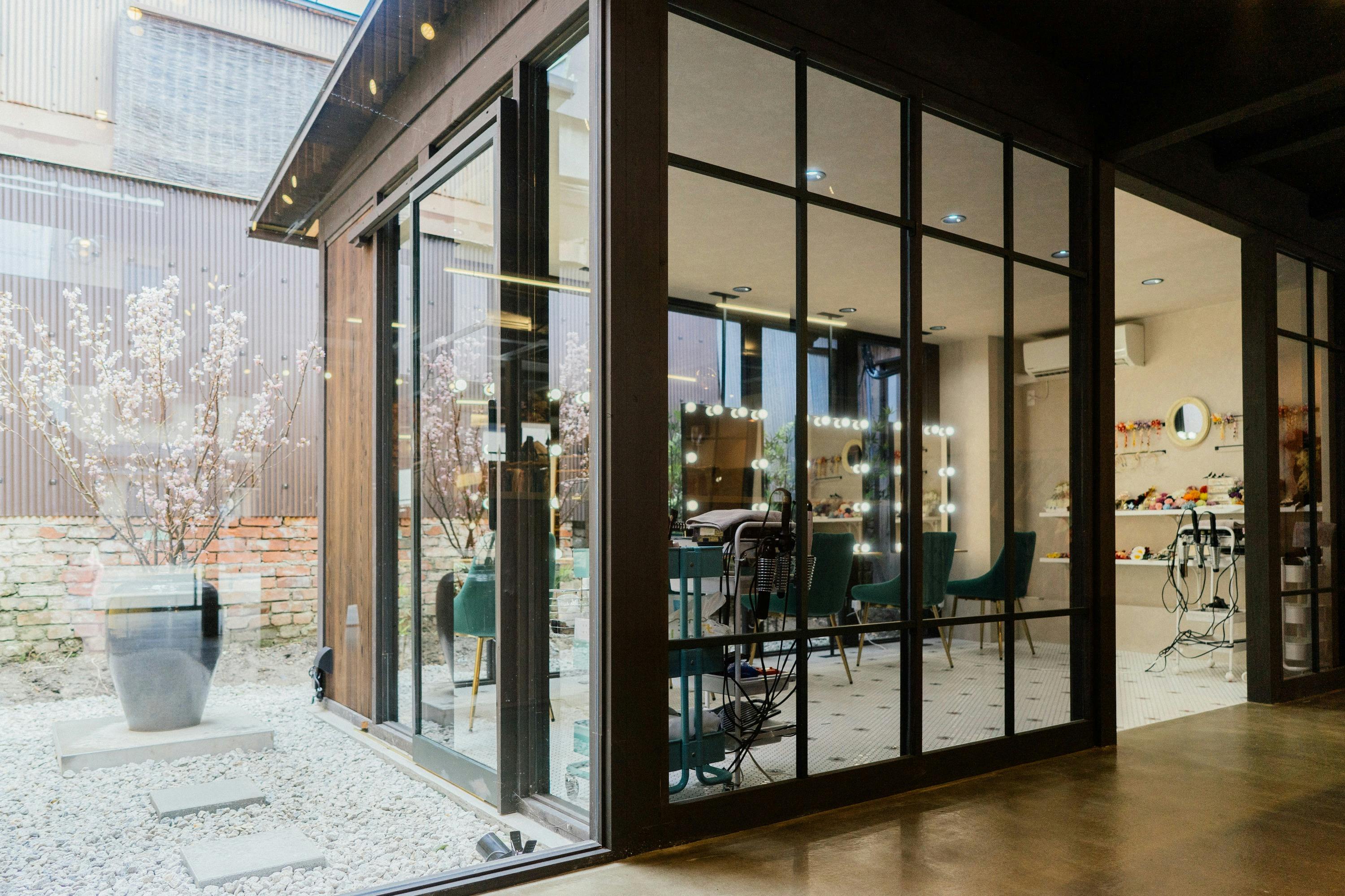 Modern salon interior with large glass windows, green chairs, and bright vanity mirrors, seen from outside.