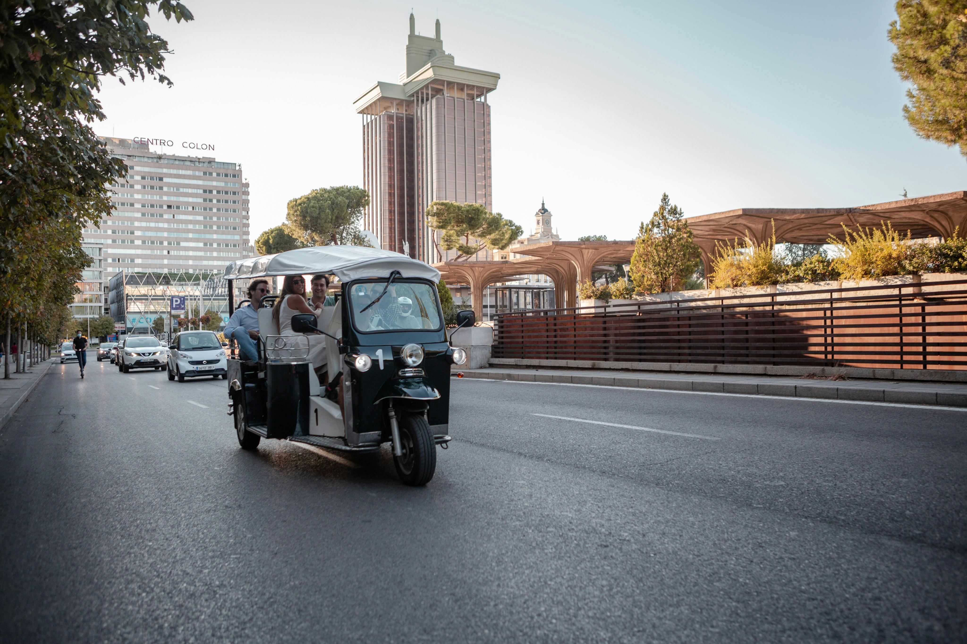 Three people ride a tuk-tuk on a city street with buildings, trees, and another vehicle in the background.