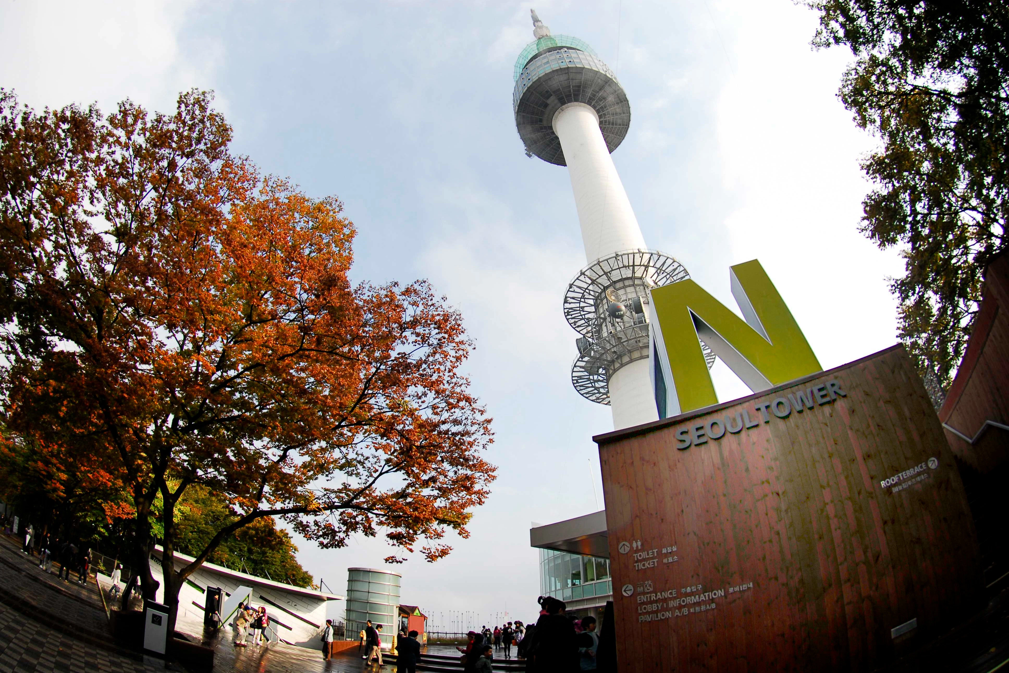 A tower with a large N at its base, surrounded by autumn-colored trees and visitors under a clear sky.