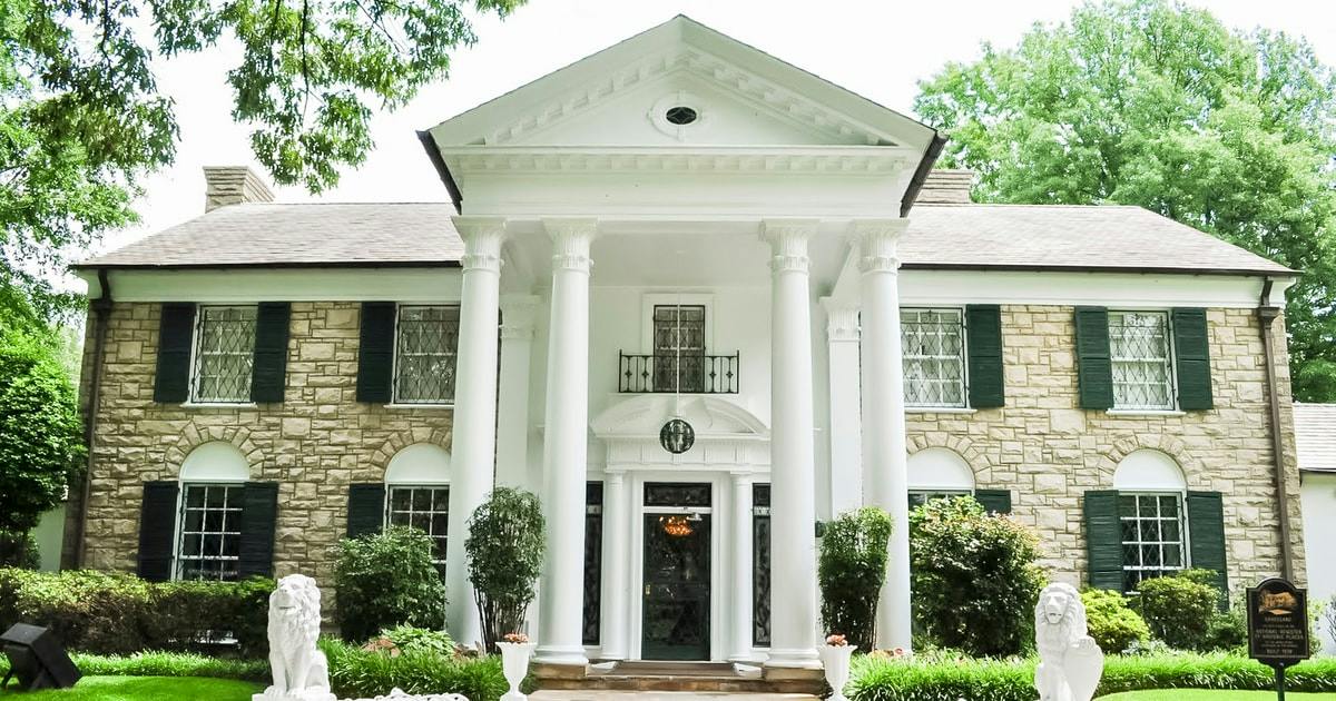 A grand white-columned entrance of a stone mansion with green shutters, surrounded by greenery and trees.