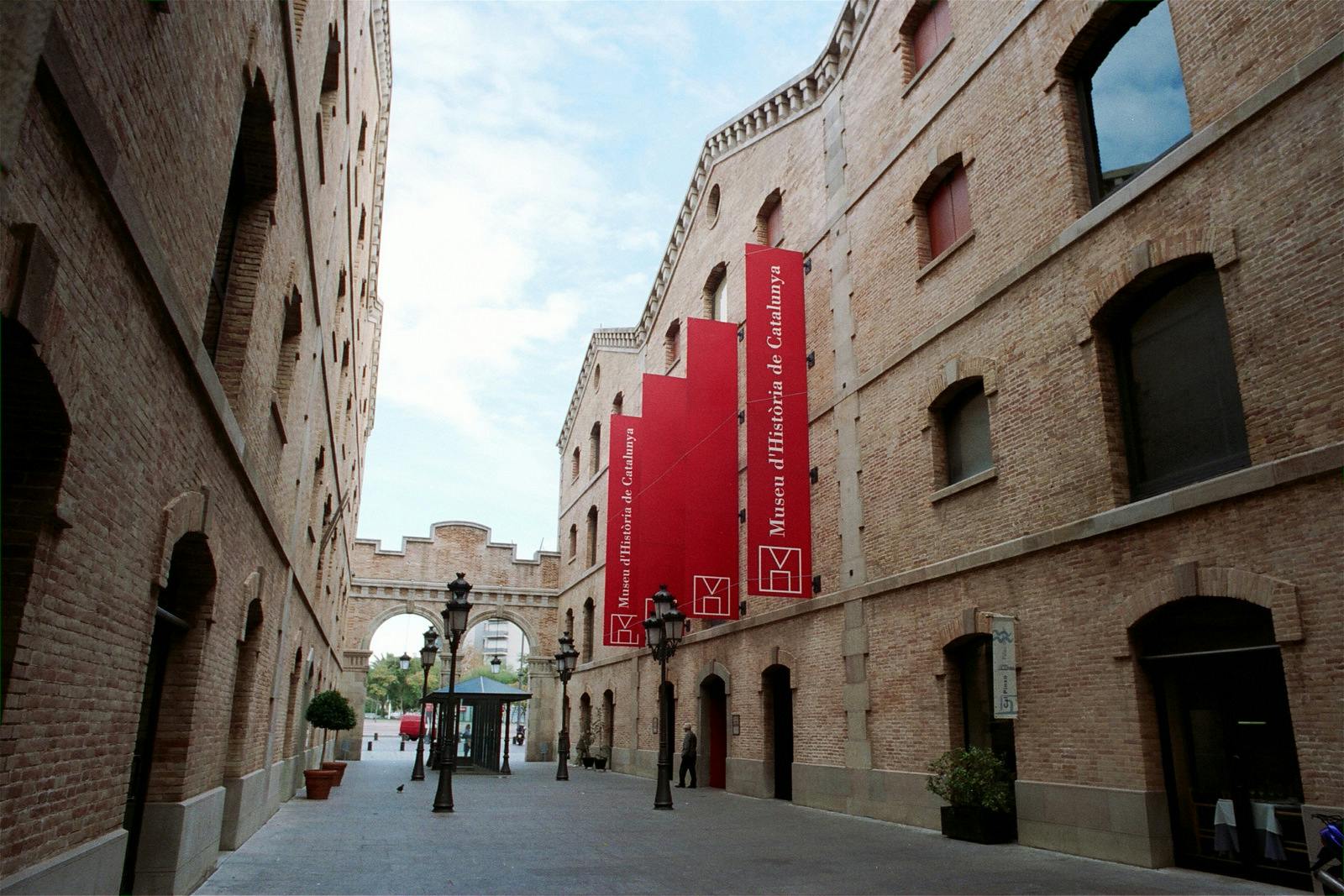Narrow street between brick buildings with red banners for Museu d'Història de Catalunya; streetlamps and potted plants visible.
