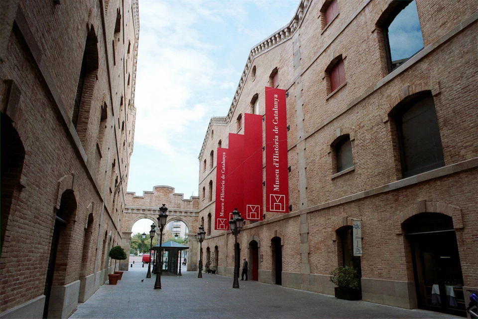Narrow street between brick buildings with red banners for Museu d'Història de Catalunya; streetlamps and potted plants visible.
