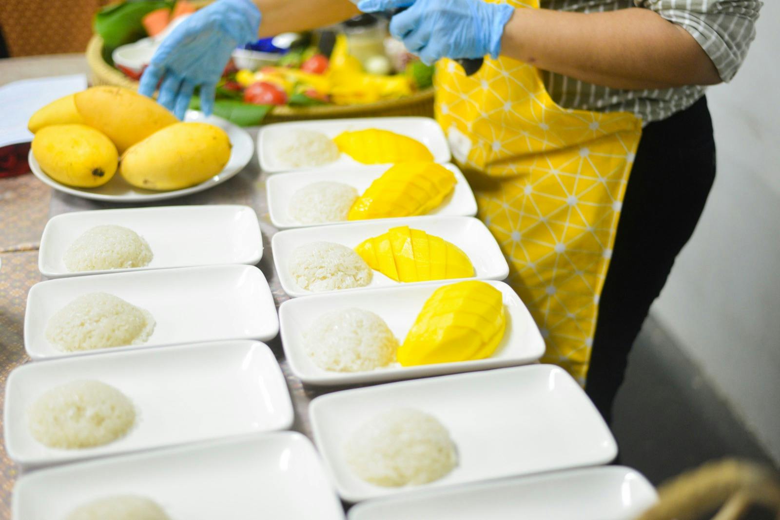 穿着黄色围裙的人准备带有糯米和芒果片的盘子。 背景中可以看到其他水果。