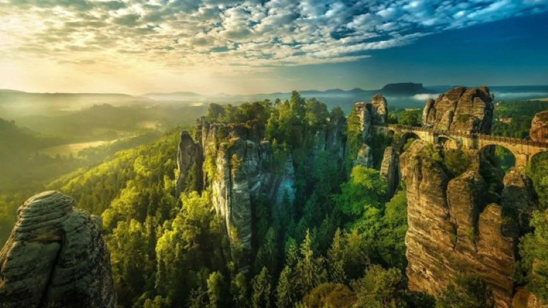 Dramatic landscape with tall rock formations surrounded by dense forest, under a partly cloudy sky with sunlight breaking through.