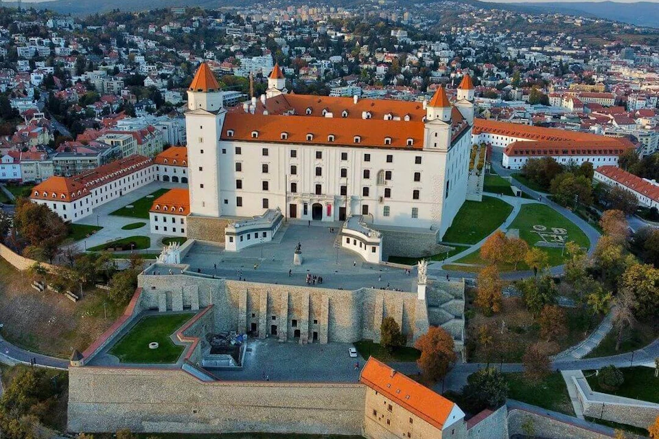 Aerial view of a large, white castle with red rooftops, surrounded by greenery and a town in the background.