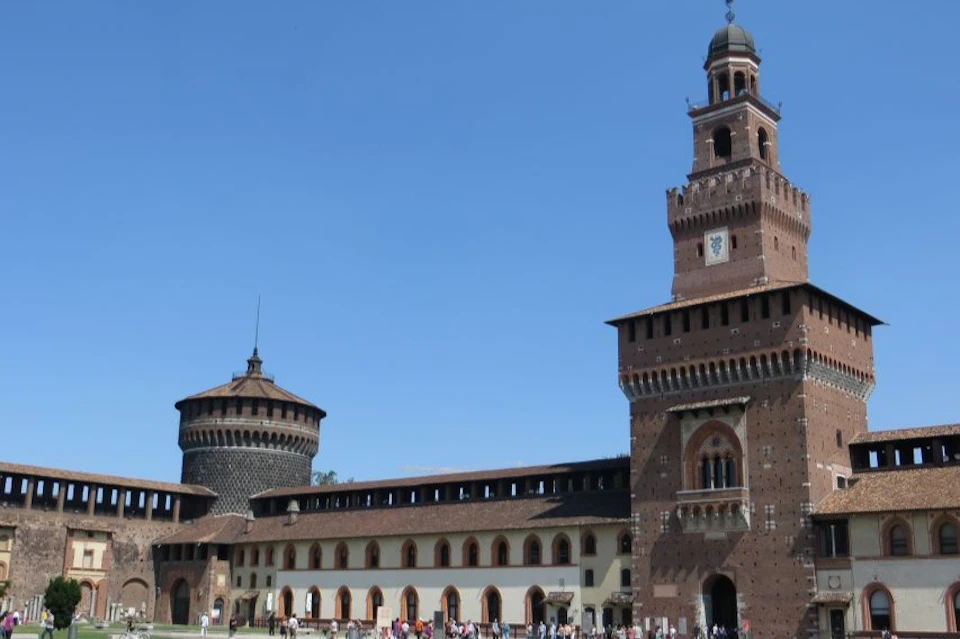 A historic castle with a tall brick tower, a round turret, arched windows, and people gathered at the base under a clear blue sky.