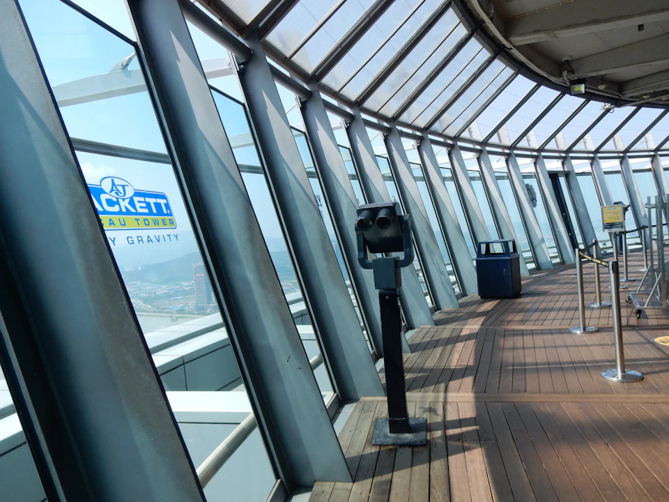 Observation deck with telescopes, large glass windows, and wooden flooring. A sign reads "AJ Hackett Macau Tower Skywalk X."
