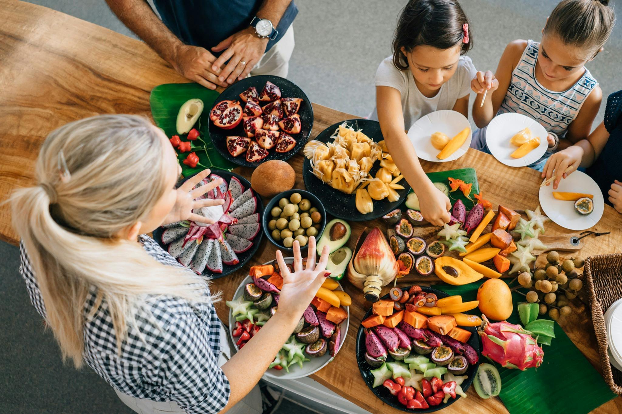 Un grup de persones al voltant d'una taula de fusta plena de fruites tropicals de colors, com fruita de drac, fruita estrellada i magrana.