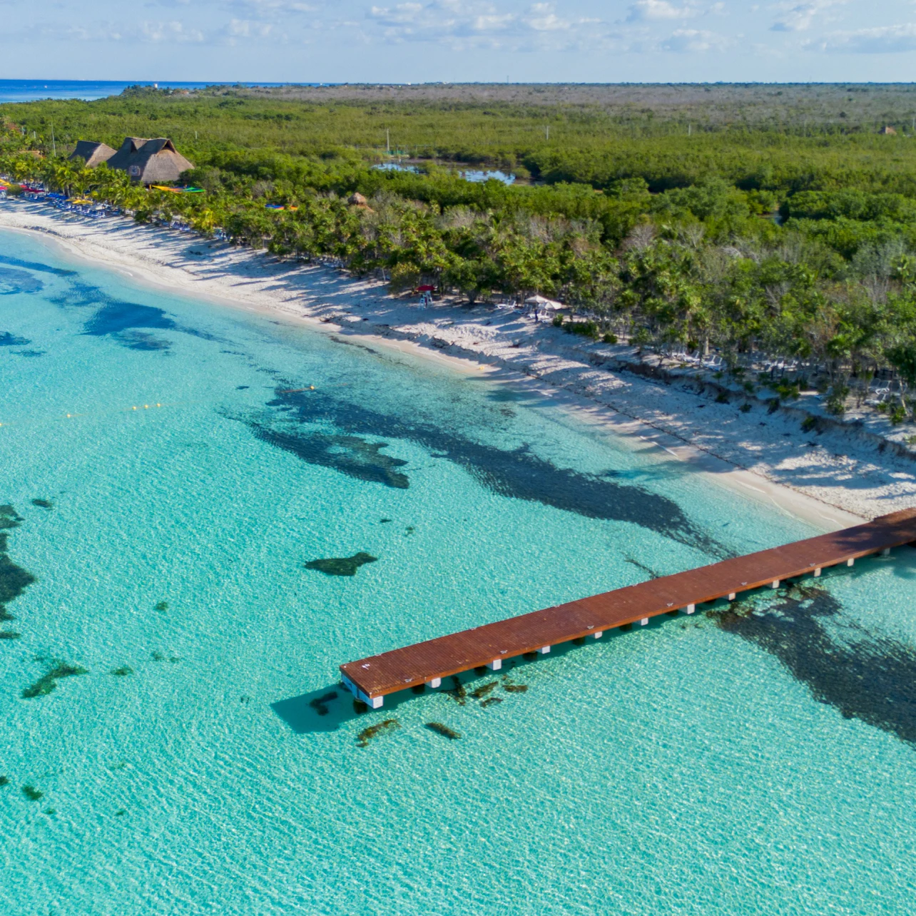 Aerial view of a wooden pier extending into clear turquoise water, with a sandy beach and dense green forest in the background.