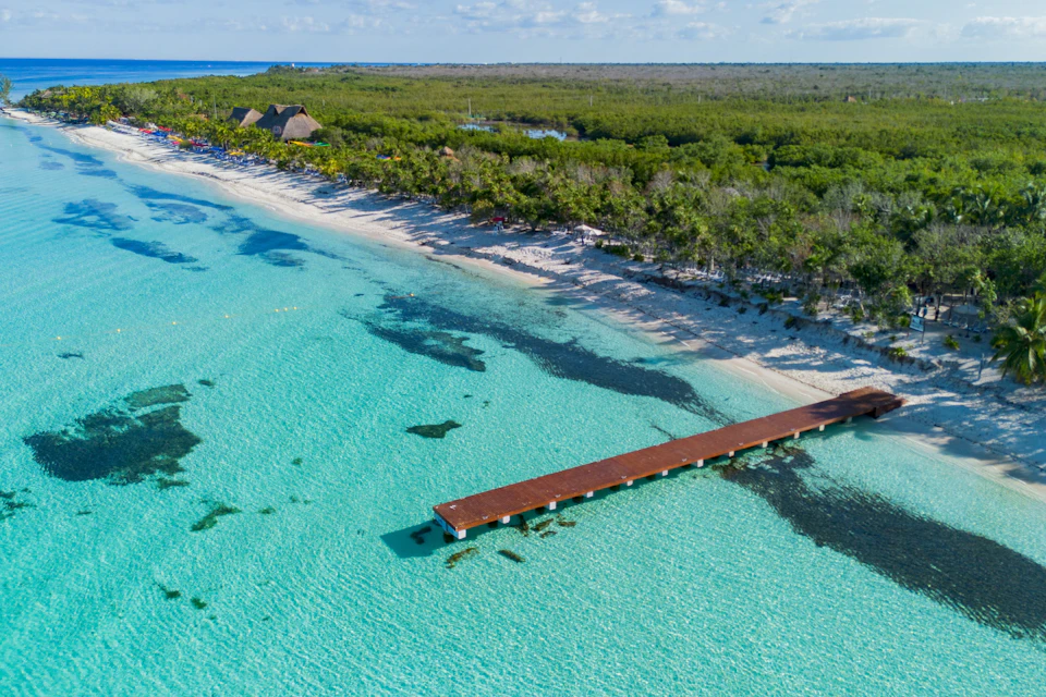Aerial view of a wooden pier extending into clear turquoise water, with a sandy beach and dense green forest in the background.