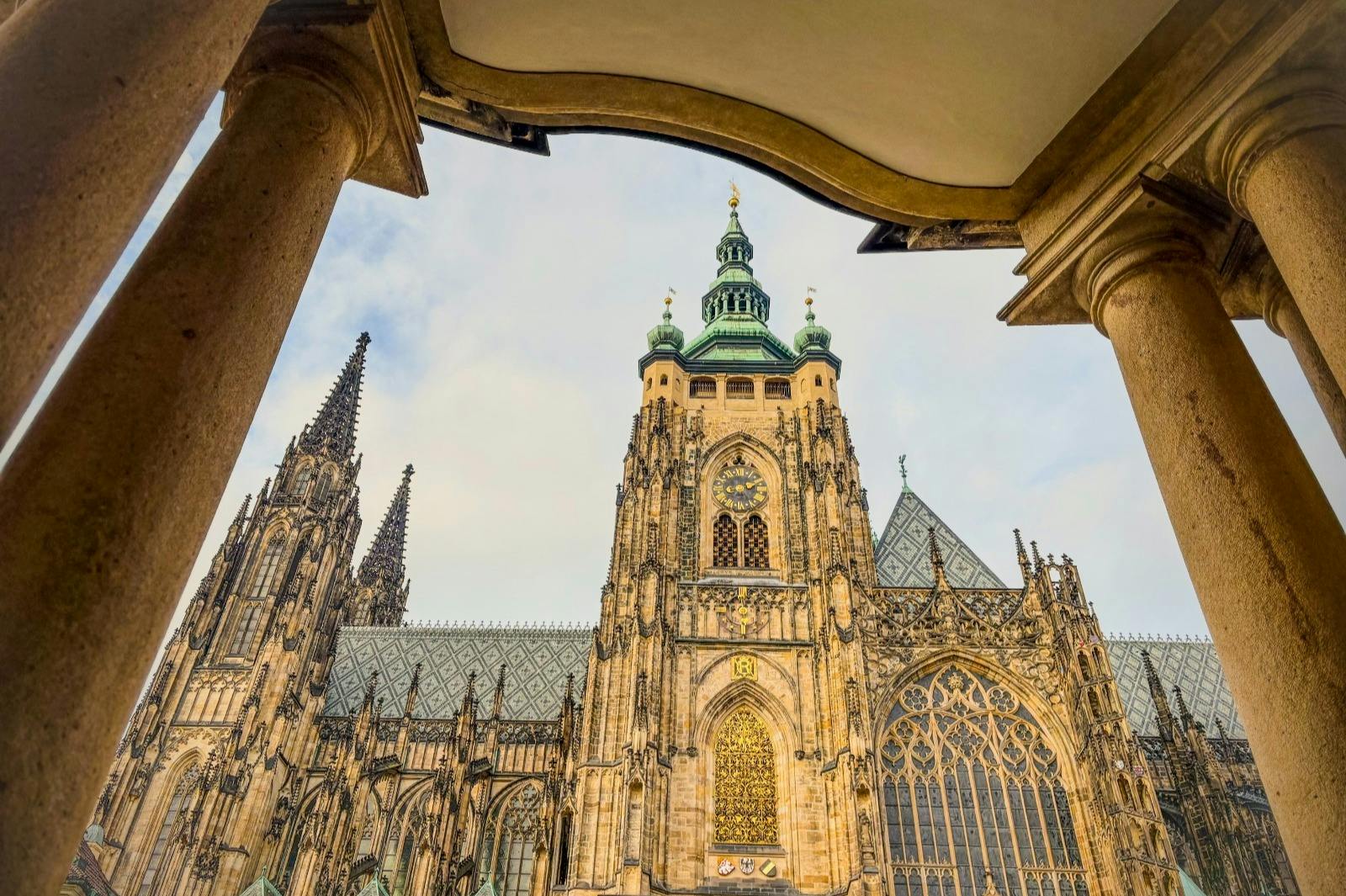 Gothic-style cathedral with intricate spires and a clock tower, viewed through an archway against a partly cloudy sky.