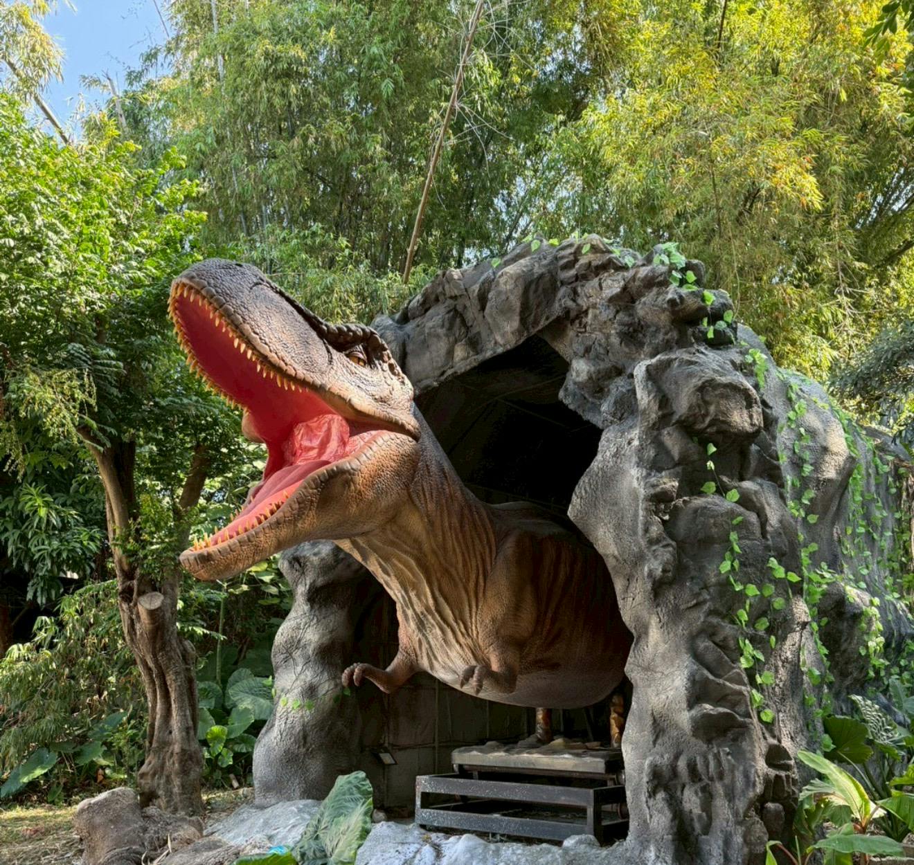 A dinosaur sculpture emerging from a rock-like enclosure in a lush, green outdoor setting.