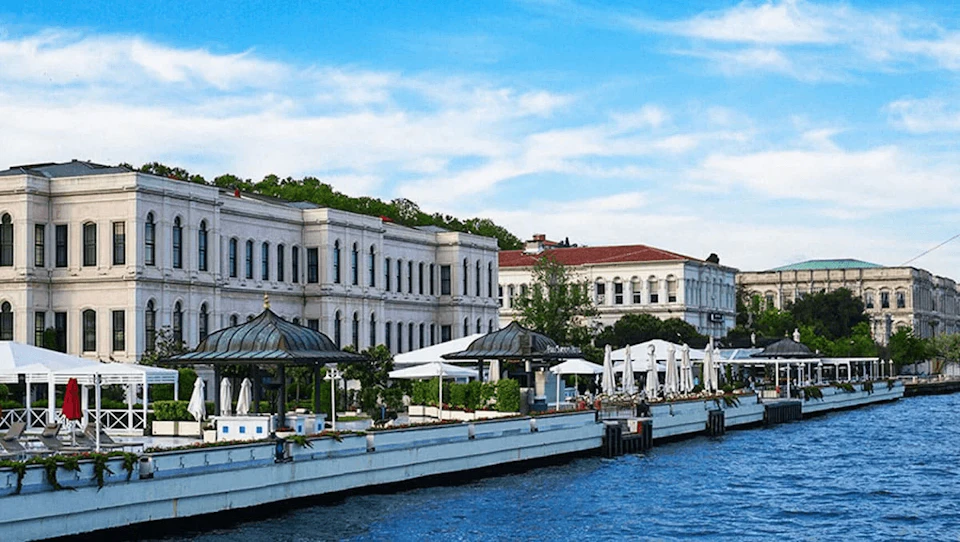 A waterfront view with historic white buildings, lush greenery, outdoor seating areas, and a blue sky with scattered clouds.