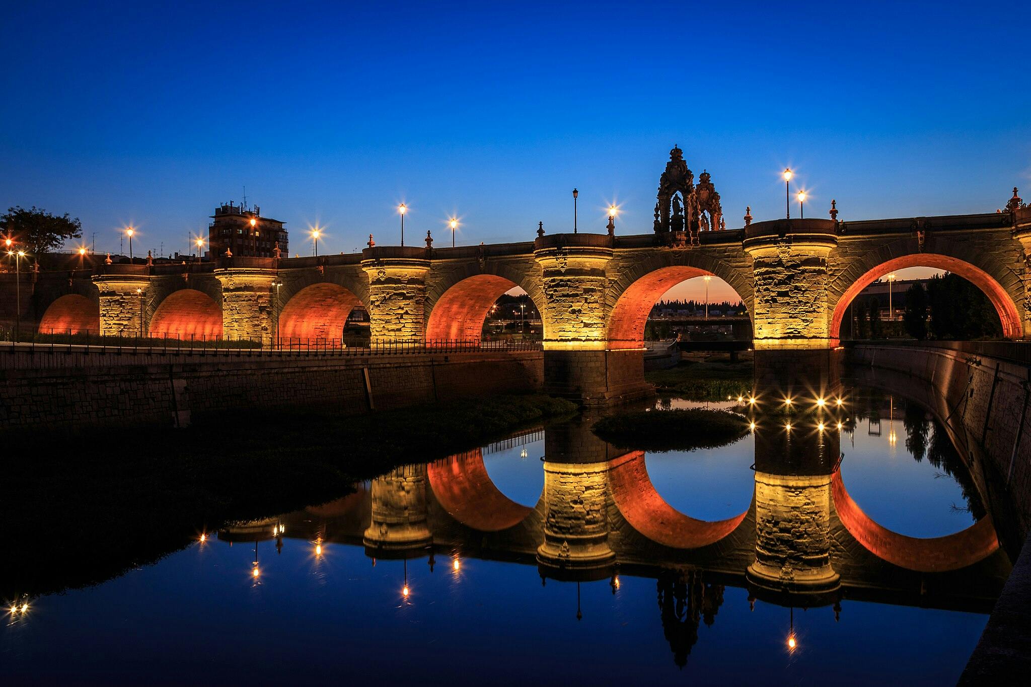 灯火通明的桥梁，拱门在黄昏时分映在平静的河流中。 灯柱和远处的建筑轮廓映衬着深蓝色的天空。