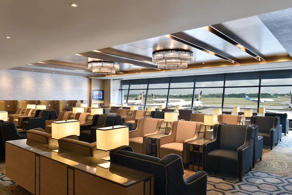 Airport lounge with beige and grey seating, modern light fixtures, and large windows showing airplanes on the runway.