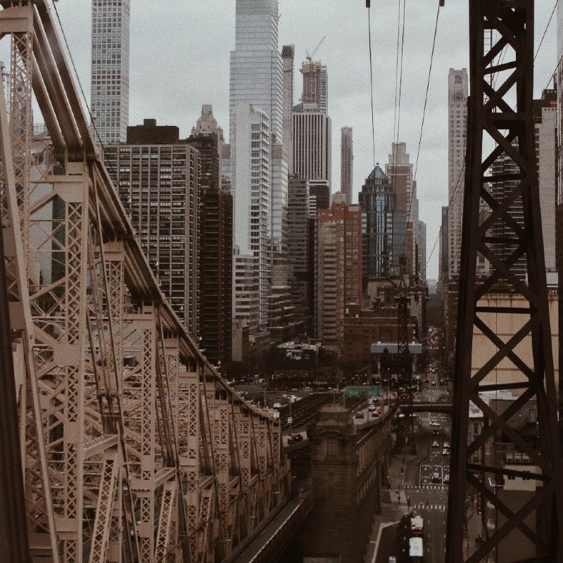 Ατσάλινη γέφυρα με αυτοκίνητα που περνούν από κάτω, με φόντο ψηλούς, πυκνοκατοικημένους ουρανοξύστες κάτω από έναν συννεφιασμένο ουρανό.