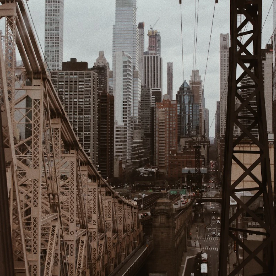 Steel bridge structure with cars passing underneath, set against a backdrop of tall, densely packed skyscrapers under an overcast sky.