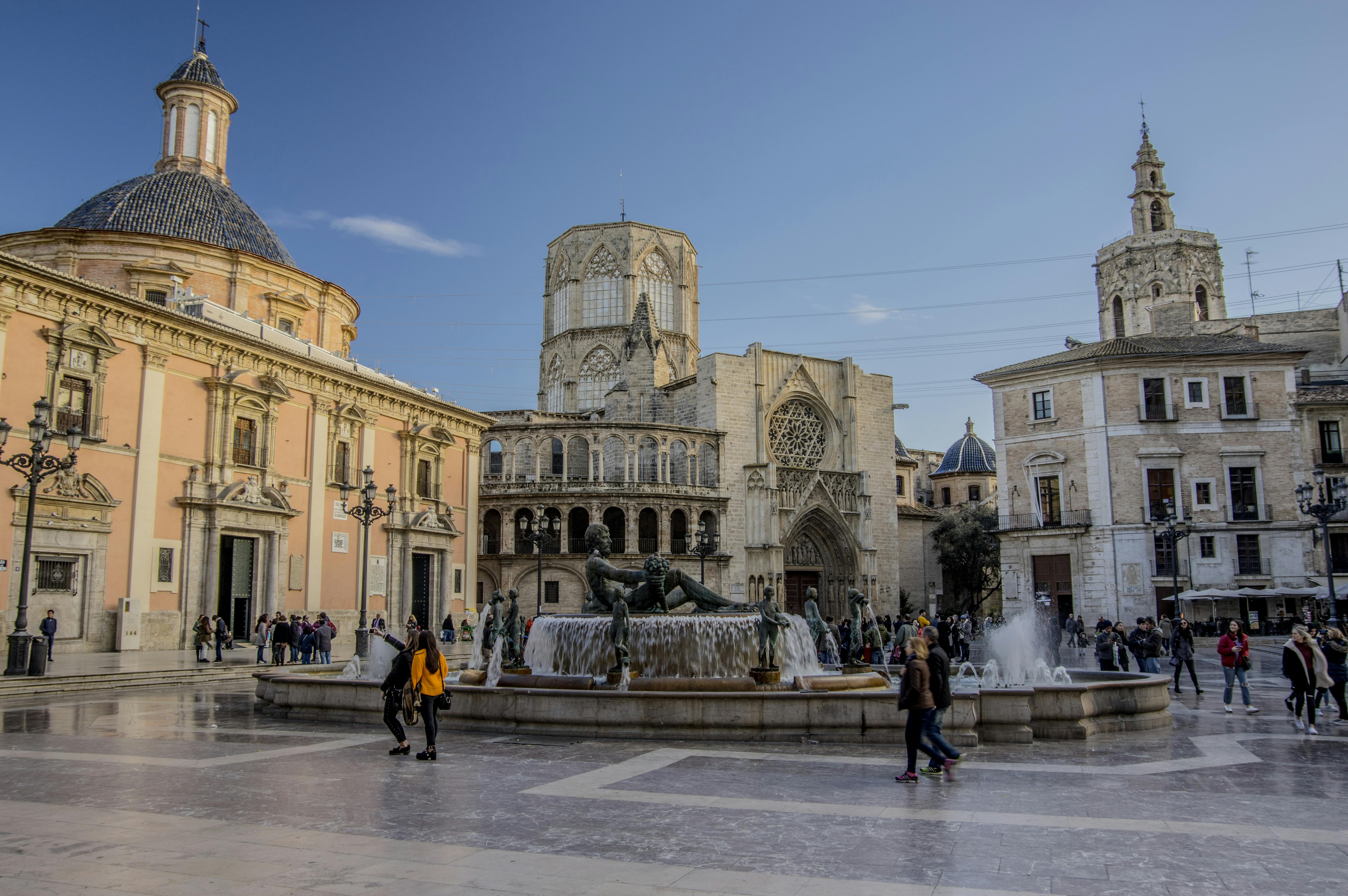 Historic square with a central fountain, surrounded by people walking and historic buildings with domes and intricate architecture.