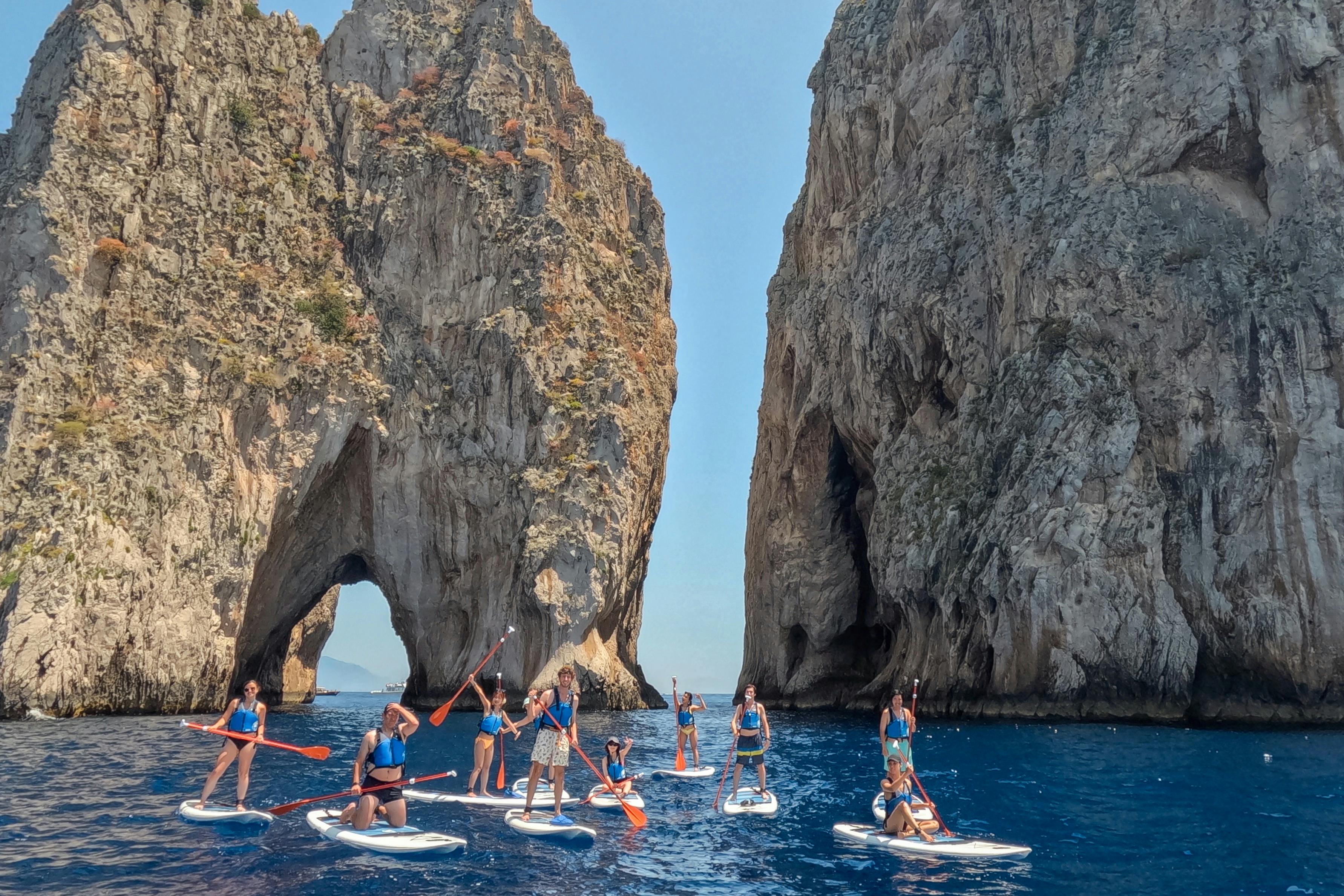 Un gruppo di persone fa paddleboarding sull'acqua blu vicino a grandi formazioni rocciose con archi naturali in una giornata di sole.