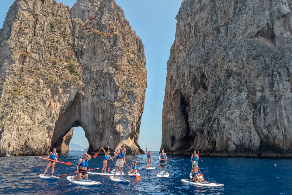 A group of people paddleboarding on blue water near large rock formations with natural arches on a sunny day.