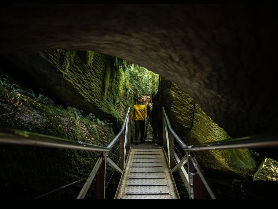 Two people walking on a metal bridge through a moss-covered rocky area with a natural overhang.