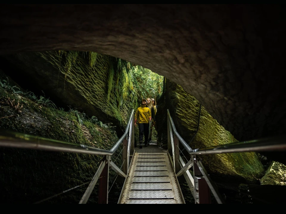 Two people walking on a metal bridge through a moss-covered rocky area with a natural overhang.