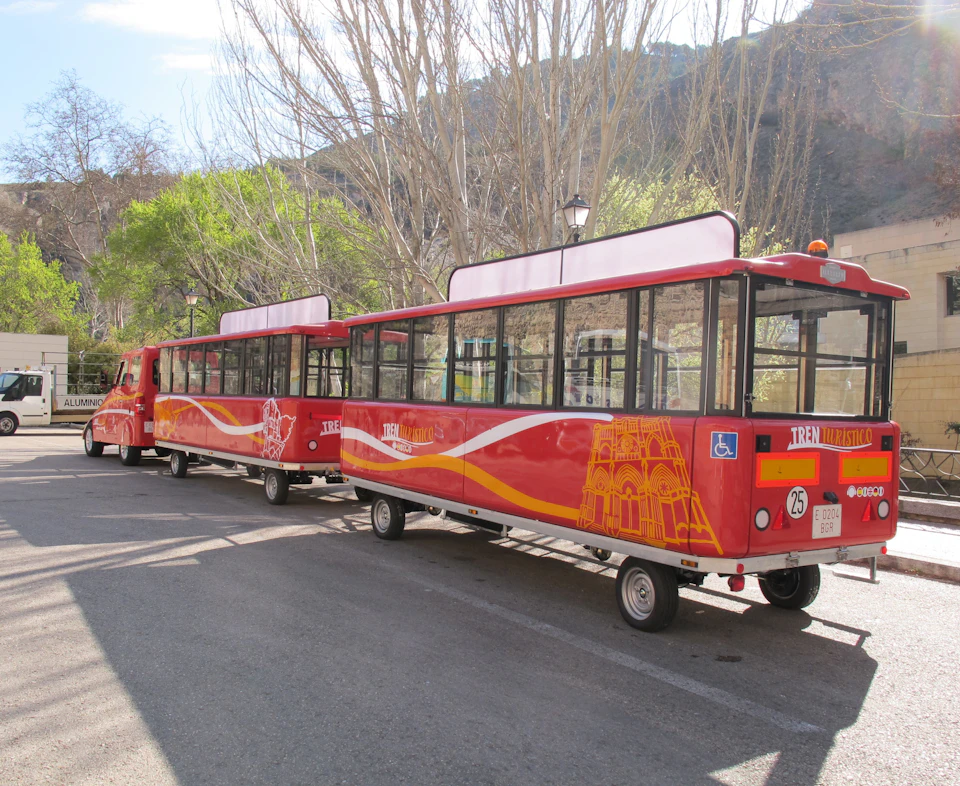 A red tourist train with two carriages parked on a street near bare trees and greenery under a clear sky.