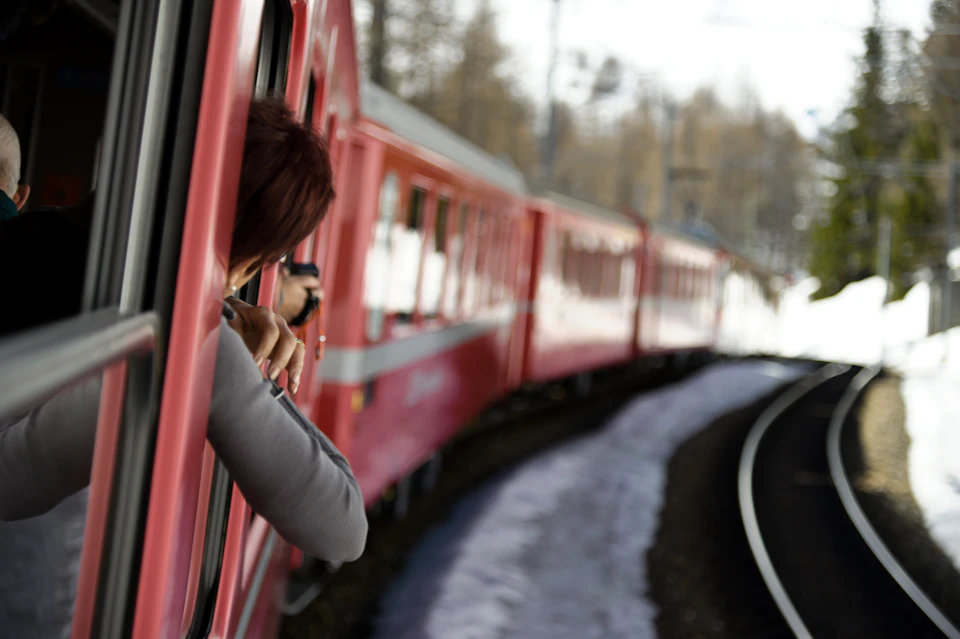 Person leaning out of a red train window, taking a photo, as the train curves along a track with snowy ground nearby.