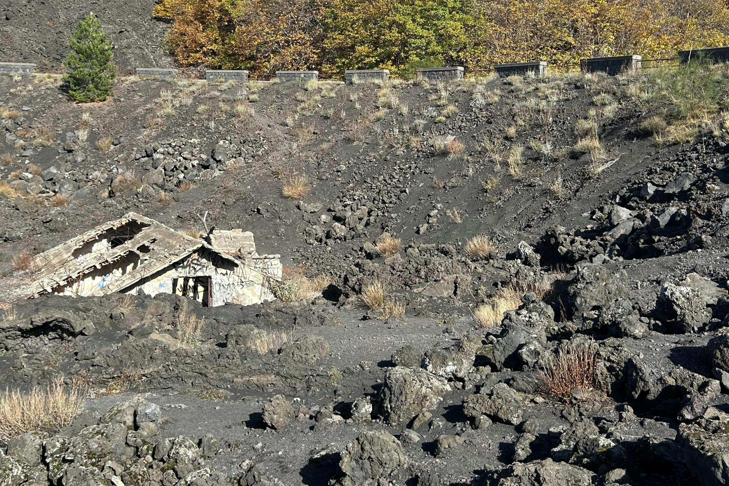Un edificio in pietra in rovina circondato da rocce vulcaniche e vegetazione rada, con degli alberi sullo sfondo sotto un cielo limpido.