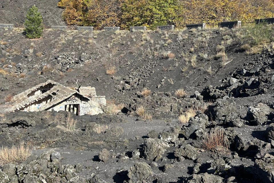 A ruined stone building surrounded by volcanic rocks and sparse vegetation, with trees in the background under clear skies.