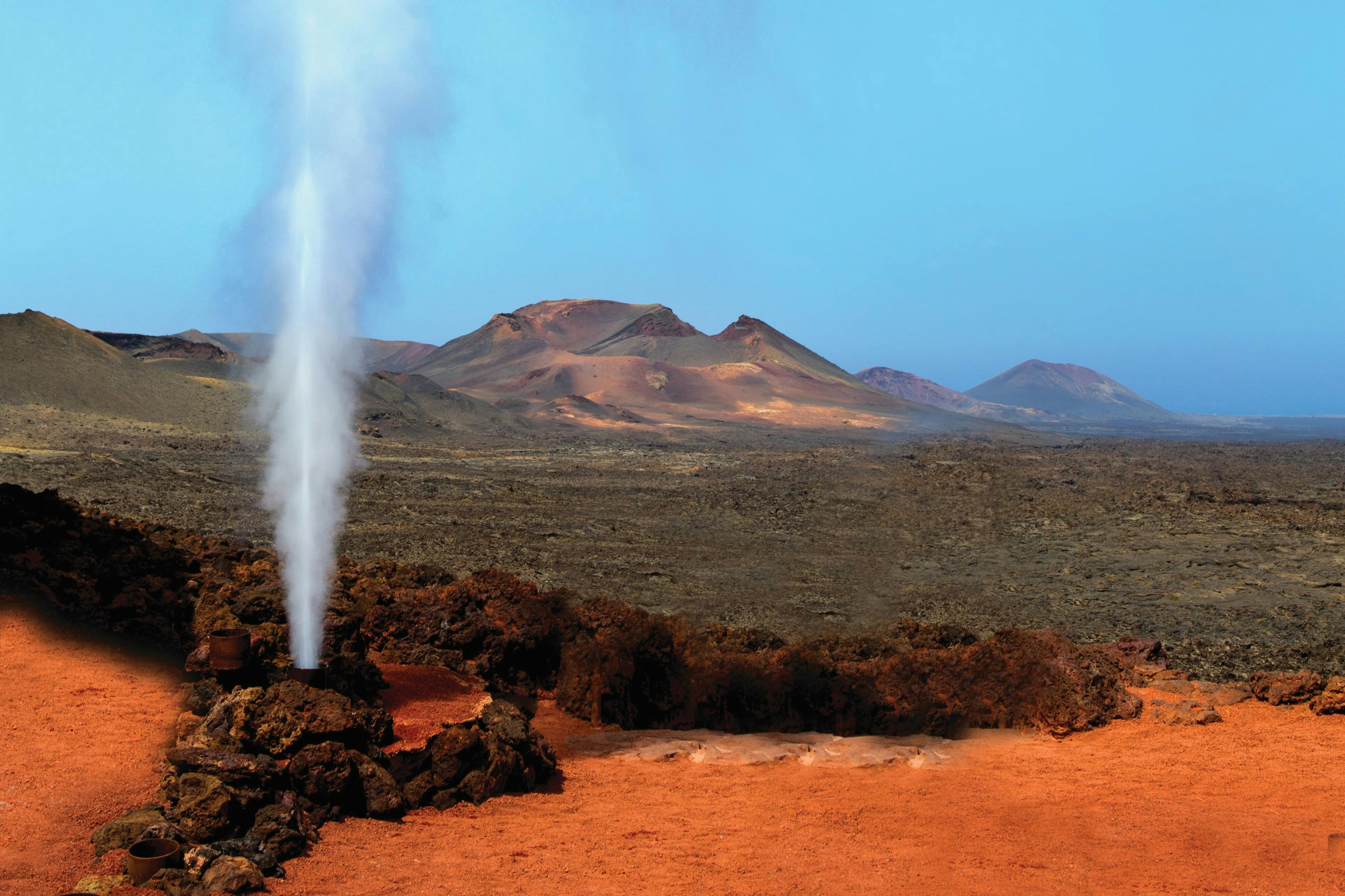 Steam geyser erupts from rocky terrain with red soil, set against a backdrop of barren, mountainous landscape under a clear blue sky.