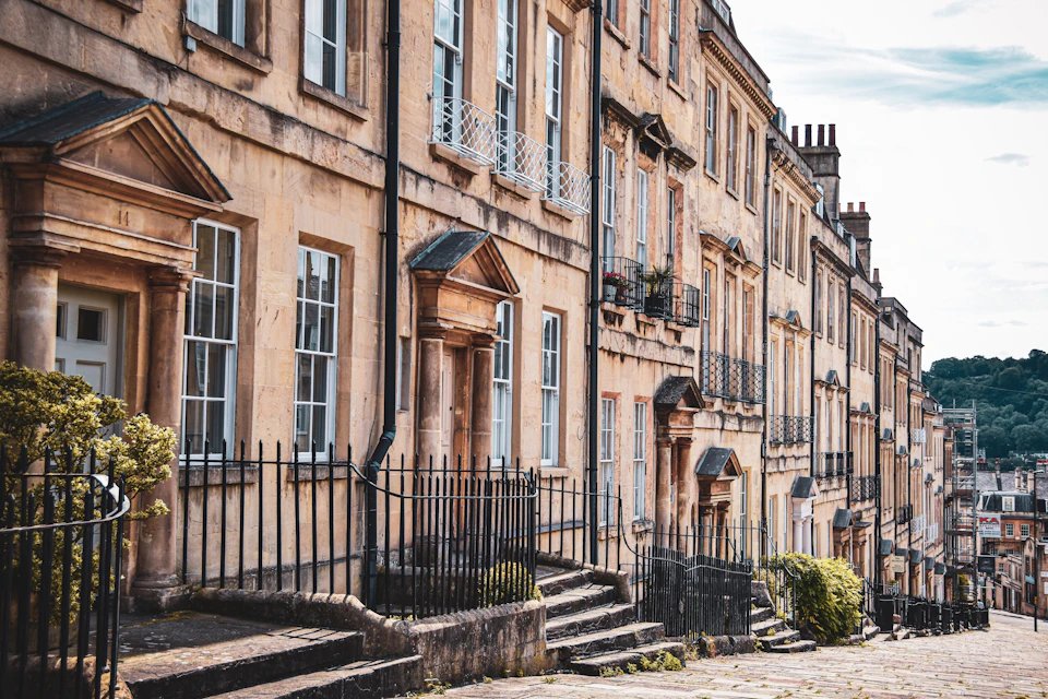 A row of historic terraced houses with stone facades, black railings, and steps leading to arched doorways on a cobblestone street.