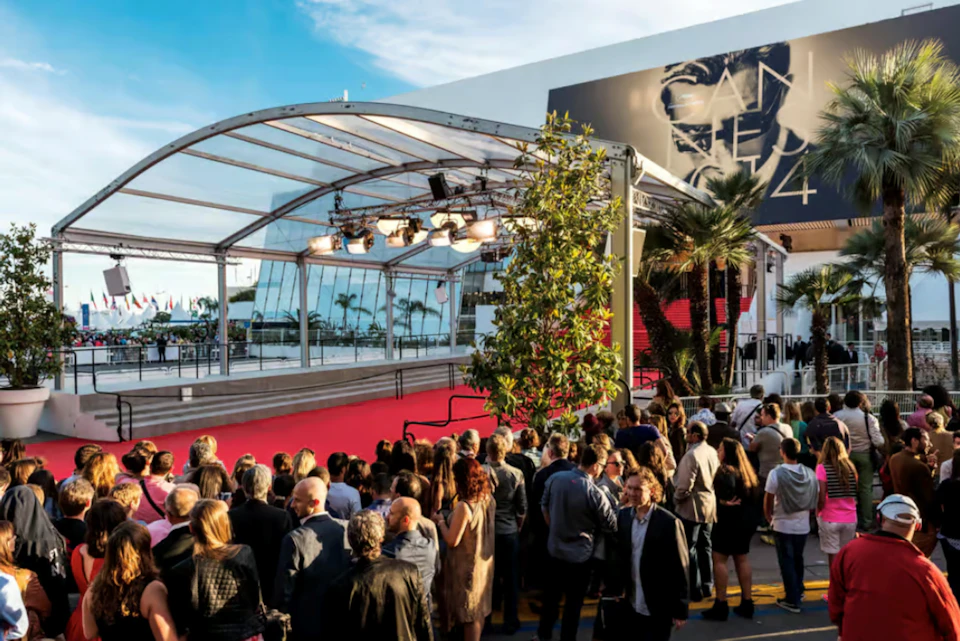 Crowd gathered outside a building with a covered red carpet area and stage lighting, under a partially clear sky.