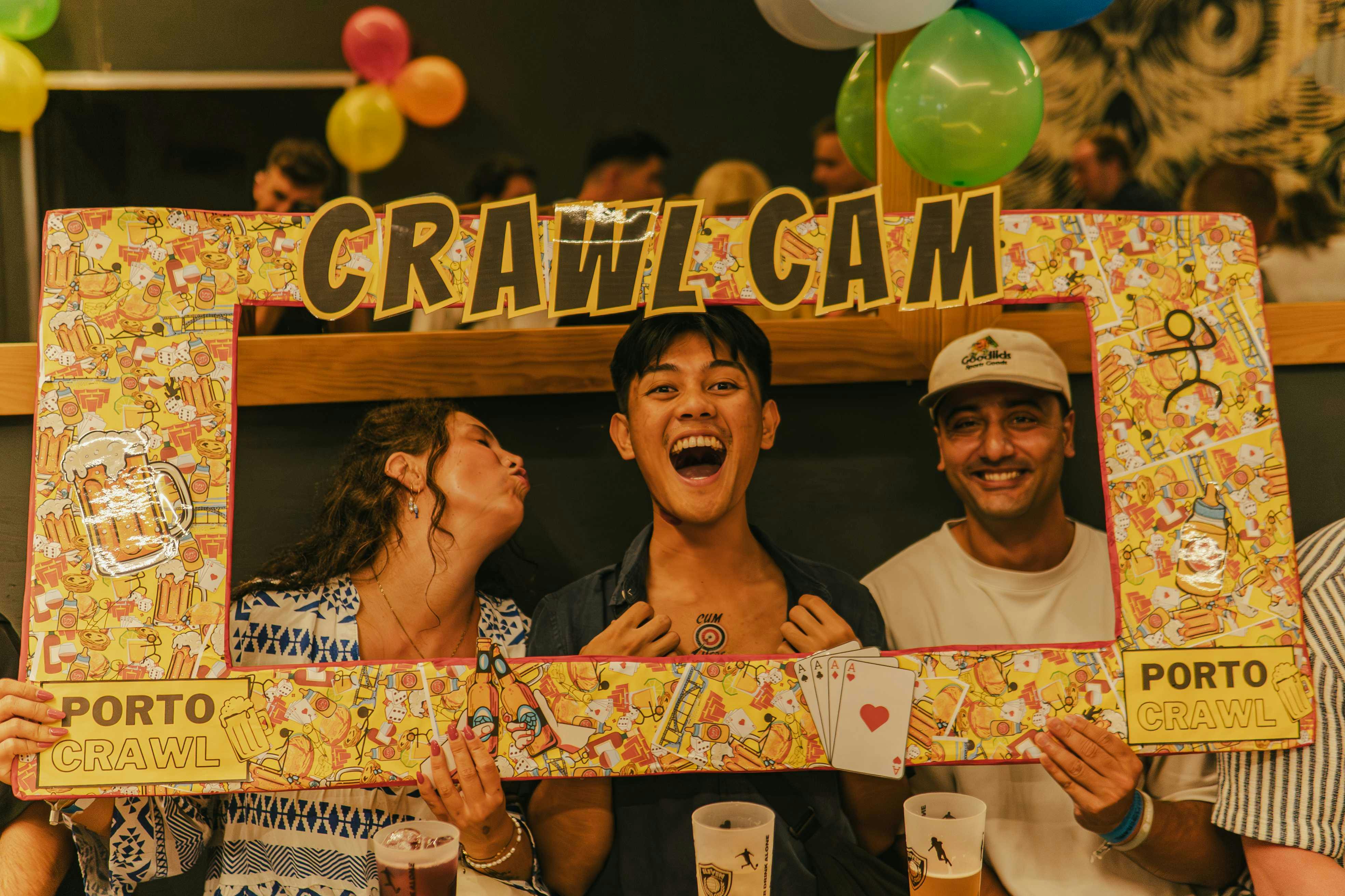 Drie mensen lachen en poseren met een kleurrijk fotolijstje met het label "Crawl Cam" in een feestelijke omgeving met ballonnen.